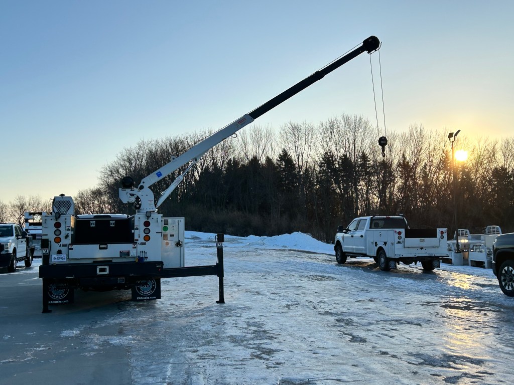 A utility truck with an extended crane arm is positioned on a snowy pavement, with trees in the background and a sunset visible.