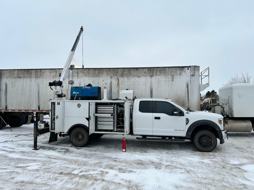 A white utility truck with a crane is parked next to a large trailer, set in a snowy outdoor environment.